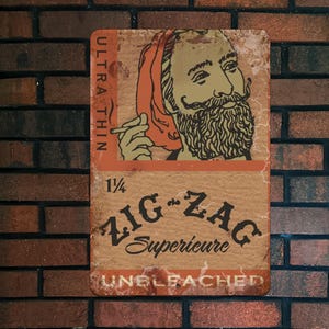 May include: A rectangular metal sign with a vintage design. The sign features an illustration of a man with a beard and mustache, holding a cigarette. The text "ULTRA THIN", "1 1/4", "ZIG-ZAG Superieure", and "UNBLEACHED" are also present.