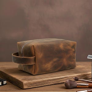 May include: A brown leather wash bag with a zip closure and a handle. The rectangular bag sits on a wooden surface. The leather has a distressed appearance. Makeup brushes and a razor are in the background.