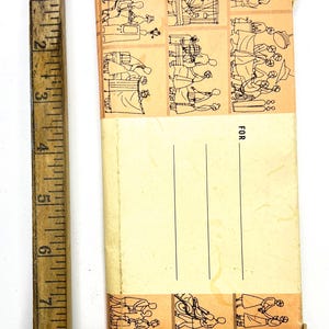 May include: A vintage notepad with a light yellow cover and a decorative border featuring small illustrations of people. The notepad has lines for writing and the word "FOR" printed on the cover. A wooden ruler is next to the notepad.
