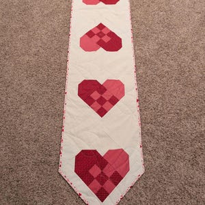 May include: A quilted table runner with a white background and four red heart designs. The hearts are made of various red fabrics and patterns. The runner has a pink and red patterned border.