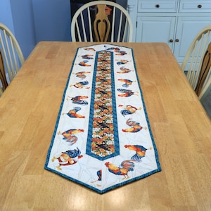 May include: A rectangular table runner with a white background and a repeating pattern of colorful roosters. The center features a floral design with a blue border. The runner is placed on a wooden table.