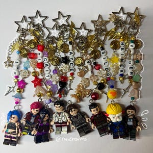 Op de afbeelding: Een verzameling sleutelhangers met Lego-achtige figuren met kleurrijk haar en outfits. Elke sleutelhanger heeft een kralenketting met ster- en goudkleurige bedels. De teksten "CAIT", "AYCE", "SILCO" en "HEIMERDER" zijn geschreven met letterkralen.
