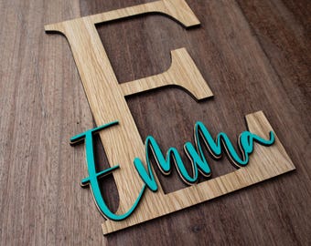 May include: Wooden sign featuring the letter "E" and the name "Emma" in a teal script font. The letter and name are layered on a wooden background, creating a 3D effect. The sign is made of wood and has a natural wood grain.