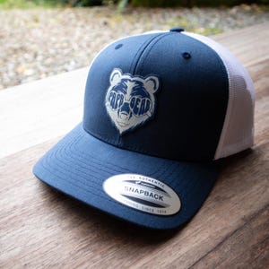 Może przedstawiać: Granatowo-biała czapka typu trucker z logo niedźwiedzia w okularach przeciwsłonecznych z napisem "PAPA BEAR". Czapka ma zakrzywiony daszek i siatkową tylną część. Jest też metka "SNAPBACK".