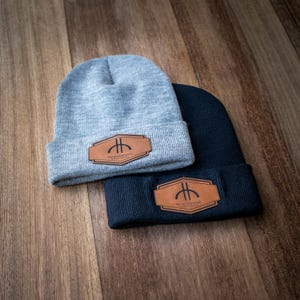 Könnte beinhalten: Zwei Strickmützen, eine graue und eine marineblaue, werden auf einer Holzoberfläche gezeigt. Jede Mütze hat einen braunen Lederaufnäher mit einem Logo. Das Logo ist ein stilisiertes Design mit Text darunter.