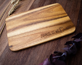 May include: A rectangular wooden cutting board with rounded corners. The board has a natural wood grain pattern and the engraved text reads "ZUHAUSE IST WO DU BIST". Decorative wheat stalks and foliage are arranged around the board.