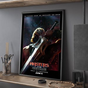 Puede incluir: Póster de película enmarcado de "Masters of the Universe" con un fondo oscuro y el texto "Witness How He Became He-Man". El póster presenta un personaje con una espada. El título de la película está en rojo y blanco, con "Only in Theaters June 5."
