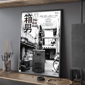 Puede incluir: Un póster en blanco y negro enmarcado titulado "The Box Man" en un entorno de habitación. El póster presenta texto japonés y una escena callejera con una estructura en forma de caja. El póster se exhibe en una estantería de madera con objetos decorativos.