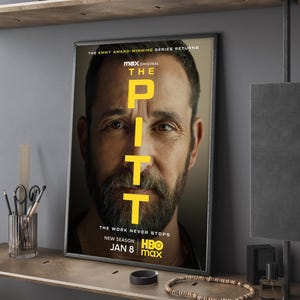 May include: Framed poster for "The Pitt" series. The poster features a close-up of a man's face with the title in large yellow letters. Text at the bottom reads "The Work Never Stops" and "New Season Jan 8 HBO Max".
