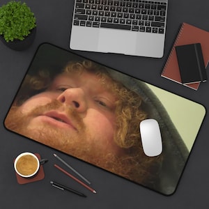 May include: A rectangular mouse mat with a close-up image of a person with red hair and a beard. The mouse mat is black with a black border. A white computer mouse is on the mat. A laptop, a notebook, and a cup of coffee are also visible.