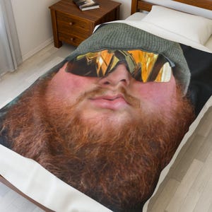 May include: A bedspread featuring a close-up of a person's face with a red beard, sunglasses, and a gray beanie. The image is printed on a white bedspread, creating a unique and eye-catching design.