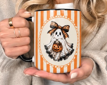 May include: A 15 oz. glossy ceramic mug with a black handle and an orange and white vertical striped pattern. The center features a circular graphic of a ghost wearing a leopard print bow and holding a jack-o'-lantern pumpkin.