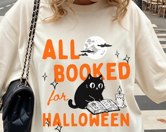 May include: An ivory t-shirt featuring a graphic print with the orange text ALL BOOKED for HALLOWEEN, a black cat reading an open book, a lit candle, a full moon, and small star accents. The shirt is made of 100% US cotton and is garment-dyed.