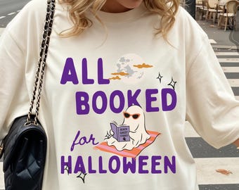May include: An ivory short-sleeve t-shirt featuring the purple text ALL BOOKED for HALLOWEEN and a graphic of a ghost wearing orange sunglasses while reading a book on a towel under a moon. The shirt is made of 100% US cotton and is garment-dyed.