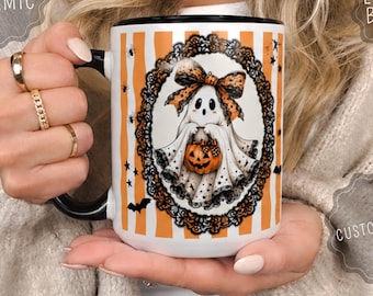 May include: A 15 oz. white ceramic mug with a black handle, featuring a vertical orange and white striped pattern with small black bats and spiders. The center displays a ghost wearing an orange bow, holding a pumpkin inside an ornate black frame.