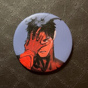 以下が含まれることがあります： 暗い髪と赤い手袋で顔を覆っている人物のアニメスタイルのイラストが描かれた丸いボタン。背景は水色です。