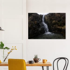 Puede incluir: Fotografía de una cascada que cae por oscuros acantilados rocosos. El agua es blanca y espumosa, contrastando con las rocas oscuras y la vegetación circundante. La imagen captura una escena de paisaje natural.