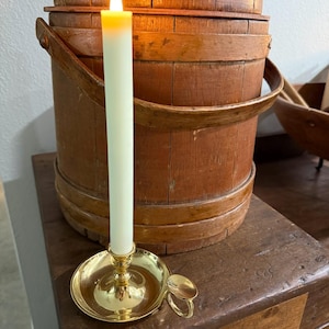 May include: A lit, pale green taper candle in a brass candlestick with a small handle. The candle sits on a wooden surface, with a rustic wooden bucket in the background. The candle flame is yellow.