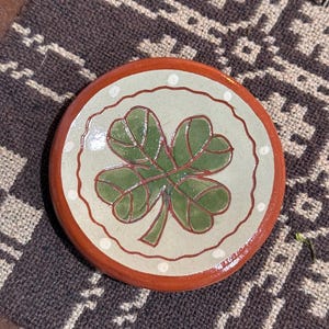 May include: A round ceramic coaster with a four-leaf clover design. The coaster has a light green center with a green and red clover, a white dotted border, and a red rim. It rests on a brown and beige patterned textile.