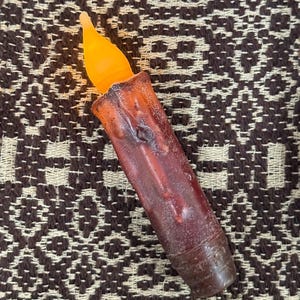 May include: A battery-operated, flameless candle with a realistic flickering orange flame. The candle body is a dark brown color with a textured, melted wax appearance. The base is a darker shade of brown.