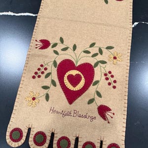 May include: Tan felt table runner with embroidered red hearts, flowers, and green leaves. The runner features the text "Heartfelt Blessings" and scalloped edges with red and green circles. A decorative home accent.
