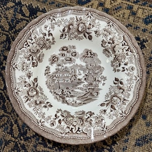 May include: A round, white and brown ceramic bowl with a floral and landscape design. The bowl features a scalloped edge and a detailed pattern of flowers, trees, and a building with a bridge. The bowl is likely vintage or antique.