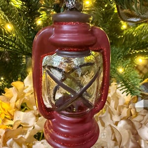 May include: A red and brown lantern-shaped Christmas ornament with glitter accents. The glass section of the lantern is clear with brown crossbars. The ornament hangs from a silver metal loop and is set against a backdrop of a Christmas tree.