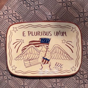 May include: Rectangular ceramic plate with a cream-colored base and a brown border. The plate features a patriotic design with an eagle holding a flag, the text "E PLURIBUS UNUM", and the year "1776".