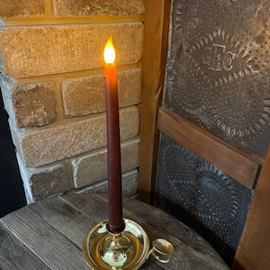 May include: A battery-operated, brown taper candle with a flickering yellow flame sits in a gold-colored candle holder. The candle holder has a small handle and is on a wooden surface. The background includes a brick wall and a decorative metal panel.