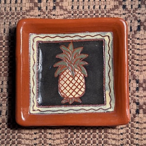 May include: A square, ceramic dish with a raised rim. The dish is a reddish-brown color. The center of the dish features a black square with a pineapple design in the center. The pineapple is yellow and white with green leaves.
