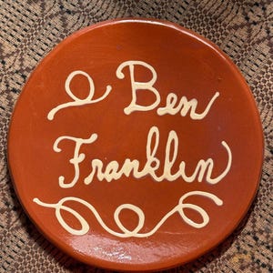 May include: A round, terracotta-colored plate with the name "Ben Franklin" written in a cream-colored, cursive font. The plate has a smooth surface and a decorative swirl design.