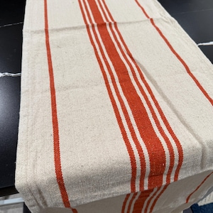 May include: Cream-colored table runner with vertical stripes in a vibrant orange-red hue. The runner is made of a textured fabric, adding visual interest. The stripes vary in width, creating a classic, farmhouse aesthetic. This textile is suitable for dining or decorative use.