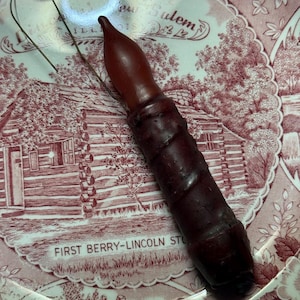 May include: A dark brown candle with a pointed top and a wire loop, resting on a red and white patterned plate. The plate features a historical scene and the text "FIRST BERRY-LINCOLN STORE".