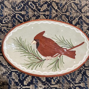 May include: Oval ceramic dish with a hand-painted cardinal bird perched on a pine branch. The bird is red with a black mask and white beak. The dish has a light green background and a brown rim.