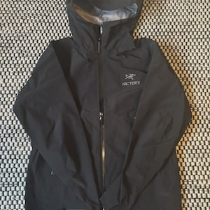 Op de afbeelding: Zwarte jas met capuchon en een lange ritssluiting. De jas heeft een capuchon en de merknaam "Arc'teryx" is zichtbaar op de borst. De jas ligt plat op een zwart-wit patroon.