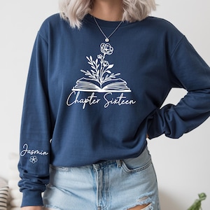May include: Navy blue long-sleeve sweatshirt with a white floral and book design. The text "Chapter Sixteen" is printed below the design. The sleeve has the name "Jasmin" and a small flower.