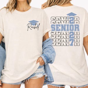 Custom Class of 2027 graduation shirt showcasing bold layered senior lettering, graduation cap artwork and personalized student name design on a relaxed fit Comfort Colors tee for graduation parties and senior photos. Blue Comfort Colors senior graduation shirt with personalized name detail, bold Senior 2027 typography and graduation themed artwork created for students celebrating their final year of high school.