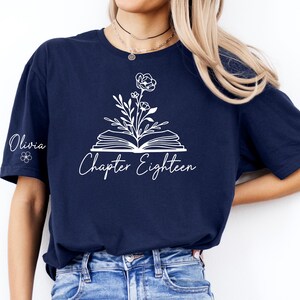 Peut inclure: T-shirt bleu marine avec un motif floral blanc sortant d'un livre ouvert. Le texte "Chapter Eighteen" est en dessous, et le nom "Olivia" est sur la manche gauche. Style décontracté.