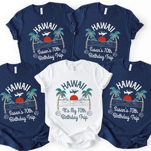 May include: Four navy blue t-shirts and one white t-shirt featuring a graphic of palm trees, a red sun, an airplane, and the text HAWAII. The navy shirts read Susan's 70th Birthday Trip, while the white shirt reads It's My 70th Birthday Trip.
