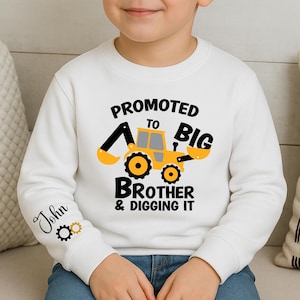 Peut inclure: Sweat-shirt blanc avec un dessin de tractopelle jaune et noir. Le texte indique "PROMU GRAND FRÈRE & DIGGING IT". La manche porte le nom "John" et des engrenages.