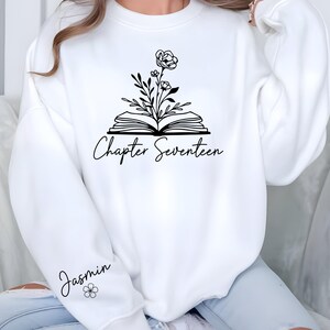 May include: White sweatshirt with a black floral design above an open book. The text "Chapter Seventeen" is below the book. The name "Jasmin" and a small flower are on the left sleeve. The sweatshirt is a casual style.
