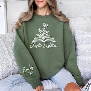 Peut inclure: Sweat-shirt vert olive avec un motif de livre floral blanc et les mots "Chapter Eighteen". Le nom "Emily" est écrit sur la manche avec un petit motif de fleur. Un haut confortable et décontracté.