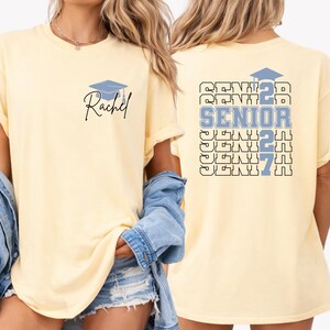 Senior year 2027 shirt featuring custom name personalization, repeating senior typography and graduation inspired graphics displayed on a blue Comfort Colors t shirt ideal for school spirit events and graduation season. Personalized graduation apparel with bold Senior 2027 design, graduation cap illustration and custom student name printed on a comfortable oversized Comfort Colors shirt for graduating high school seniors.