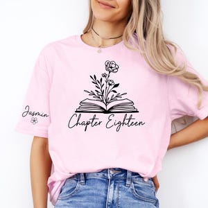 Peut inclure: T-shirt rose clair avec un motif floral noir sortant d'un livre ouvert, avec l'inscription "Chapter Eighteen" en dessous. Le nom "Jasmin" est écrit sur la manche gauche.