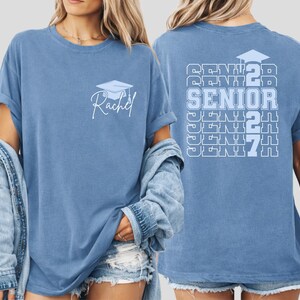 Class of 2027 senior shirt featuring stacked typography artwork, graduation themed design elements and personalized name customization on a blue vintage style Comfort Colors t shirt for memorable celebrations. Custom Class of 2027 senior shirt with repeating senior text design and personalized name graphic displayed on a vintage style blue Comfort Colors tee for graduation celebrations.