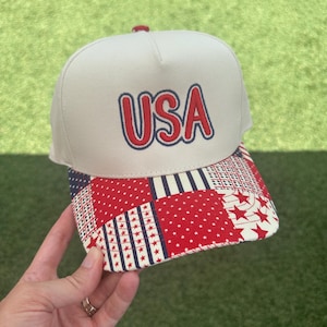 May include: A beige baseball cap with the word "USA" embroidered in red with a blue outline. The brim features a patchwork design with red stars, white stripes, and blue accents. The cap is held by a hand against a green background.