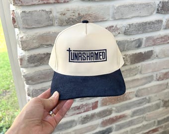 May include: A baseball cap with a cream-colored crown and a navy blue brim. The cap features the word "UNASHAMED" in navy blue block letters with a cross symbol above the text. The cap has a small navy blue button on top.
