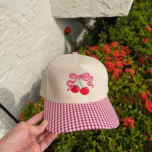 May include: A beige baseball cap featuring an embroidered design of two red cherries connected by a pink ribbon bow. The cap has a structured front panel and a flat brim with a red and white gingham checkered pattern across the entire brim surface.