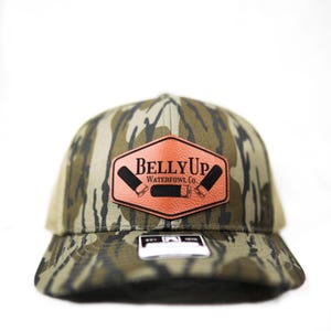 May include: Camouflage baseball cap with a brown leather patch. The patch features the text "BELLY UP WATERFOWL CO." and illustrations of shotgun shells. The cap has a curved brim and a mesh back.