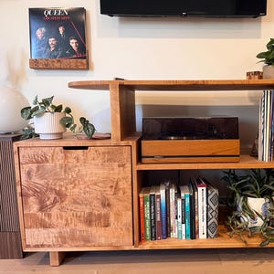 Può includere: Una console multimediale in legno con giradischi, mensole e un mobile. La console è realizzata in legno chiaro e presenta un giradischi sulla mensola centrale, con un mobile sul lato sinistro. Le mensole contengono libri, piante e un album.